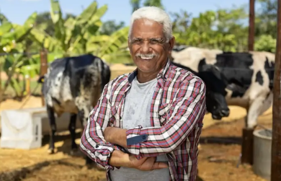 Homem de braços cruzados. No fundo há um pasto com algumas vacas