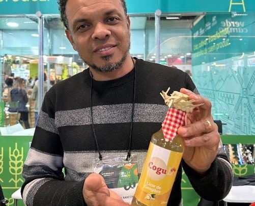 homem de barba sorrindo, segurando com as duas mãos uma garrafa de licor na cor amarela e ornamentada com chapéu de palha