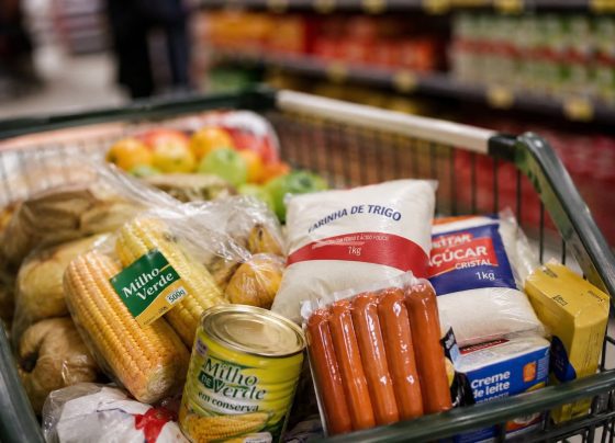 Imagem mostra carrinho de supermercado com comidas típicas de festa junina, como milho, salsicha e açúcar